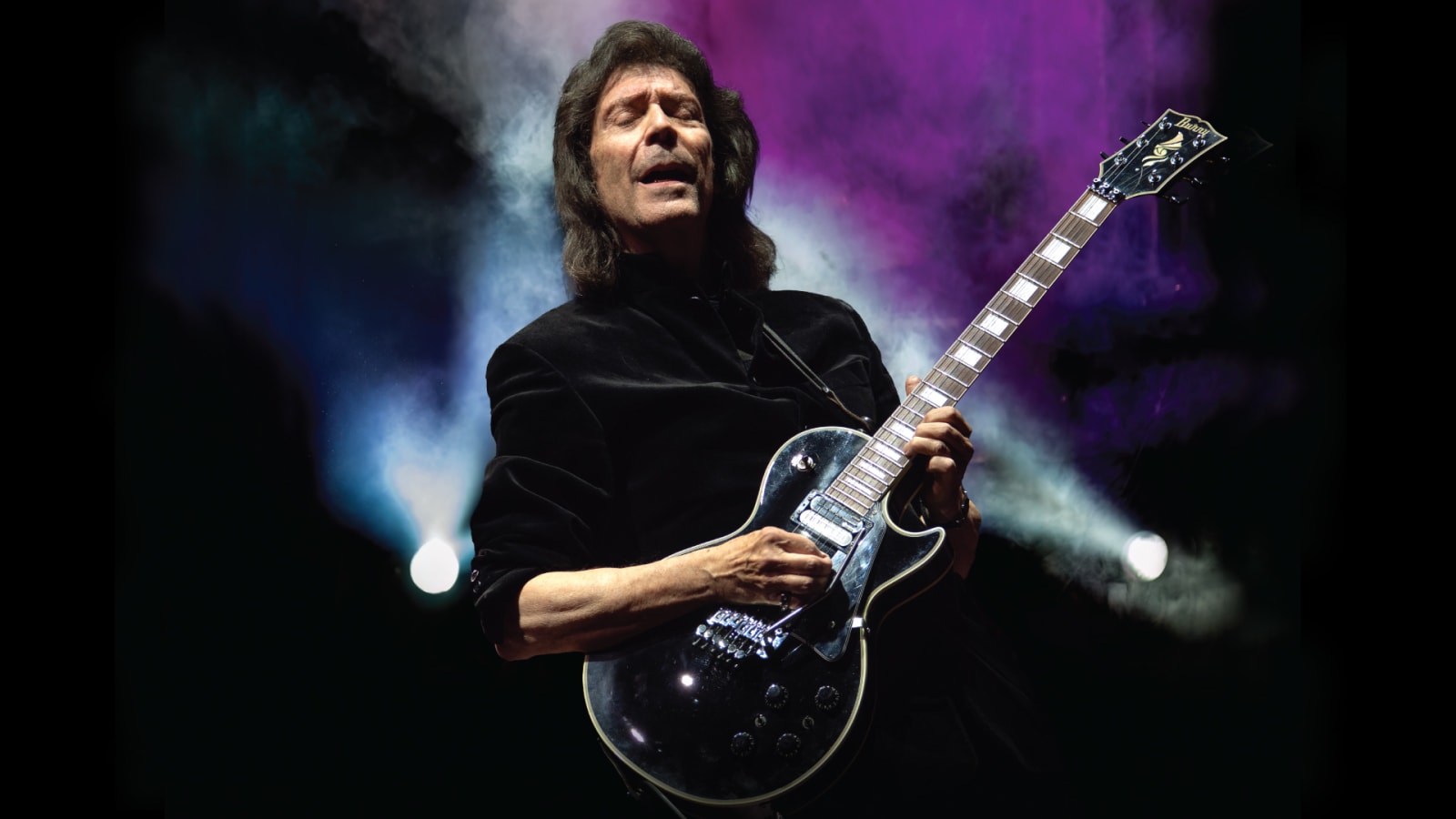 A guitarist in a black jacket plays a dark electric guitar on a dimly lit stage with beams of white and purple light through haze.