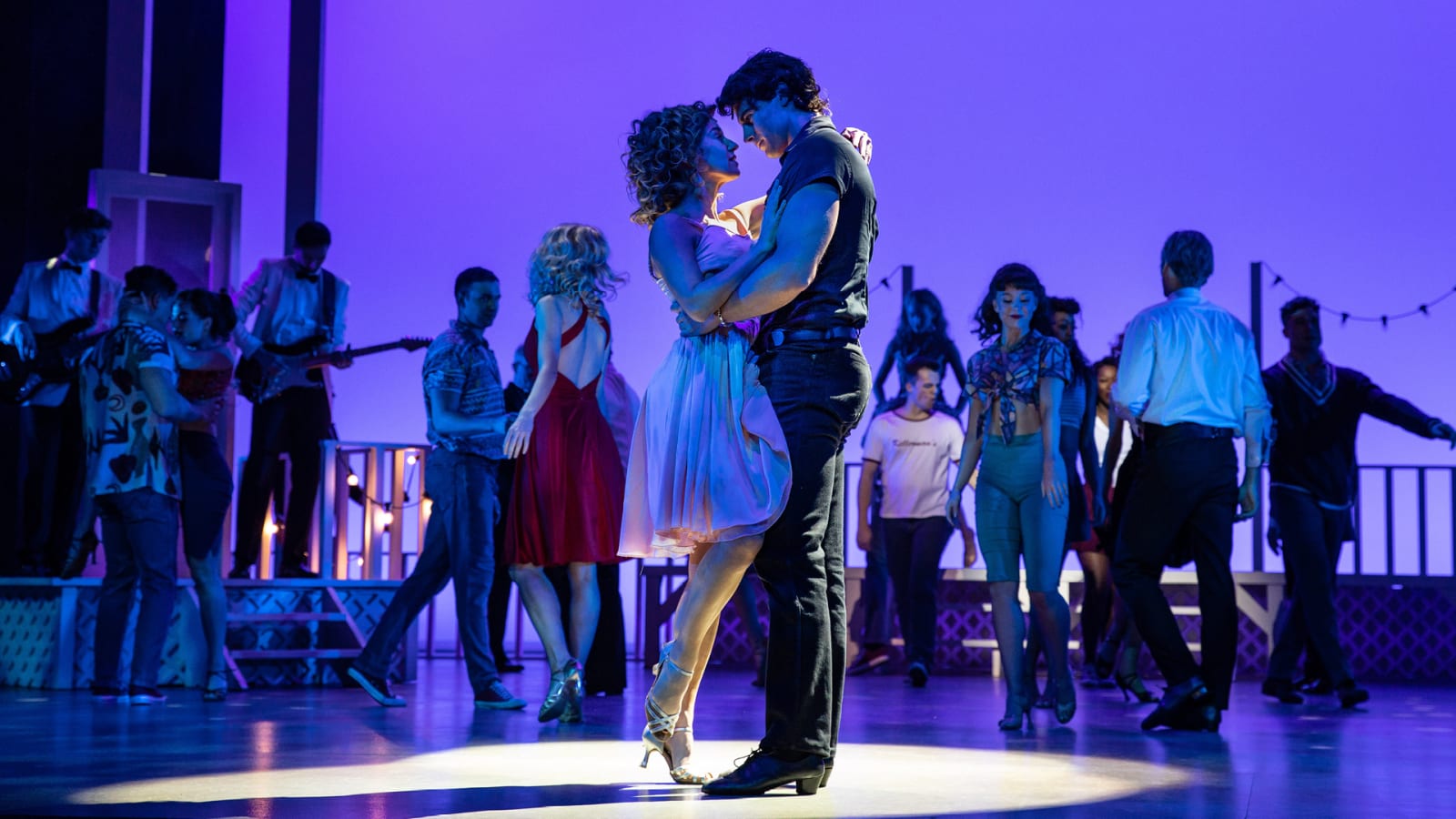 Two performers in a close embrace under a purple spotlight with an ensemble cast on a theatrical stage.