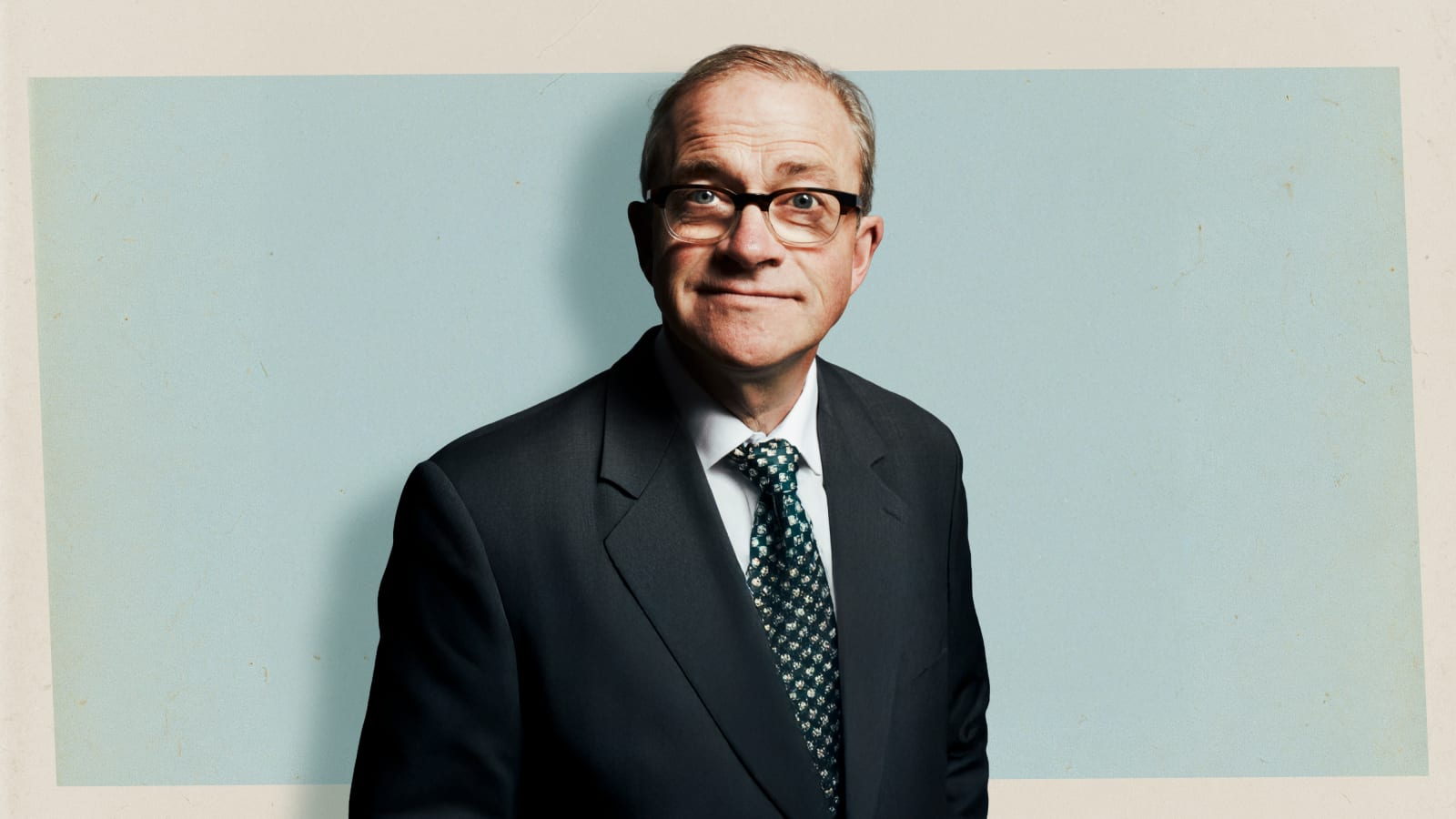 A man in a dark suit and patterned green tie, wearing black-rimmed glasses, poses with a wry half-smile against a pale blue-grey background with a cream border.