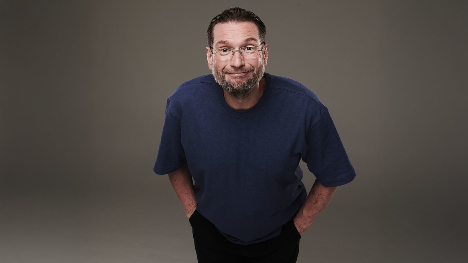 A bearded man in clear-framed glasses and a navy T-shirt leans forward against a plain grey-brown studio background.
