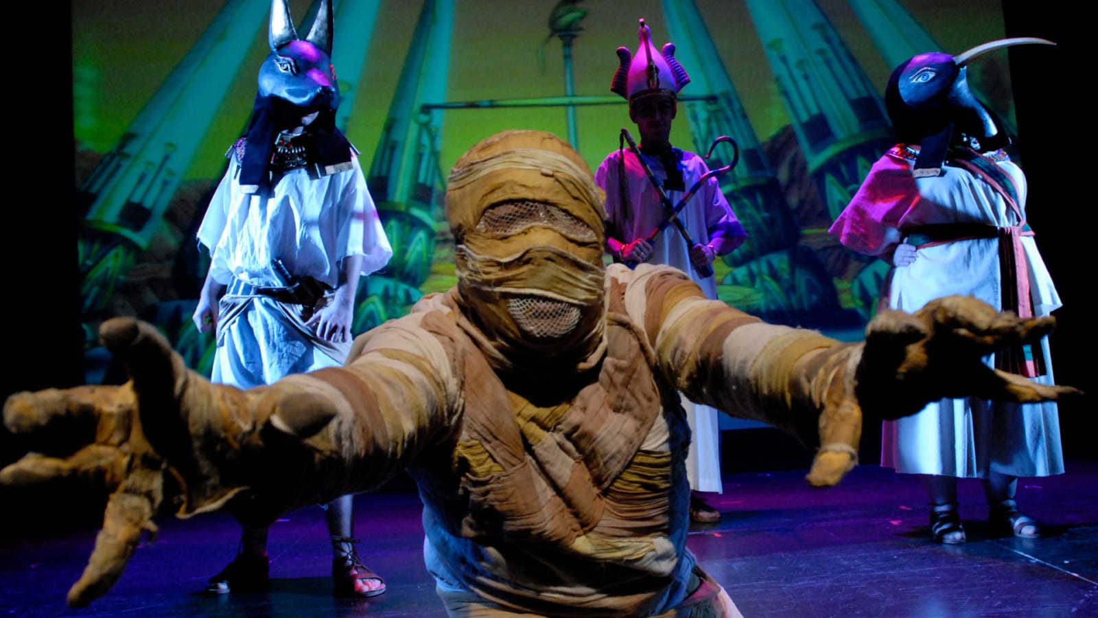 A performer wrapped as a mummy reaches forward, with animal-headed figures and a pharaoh-costumed performer behind under blue and purple light.