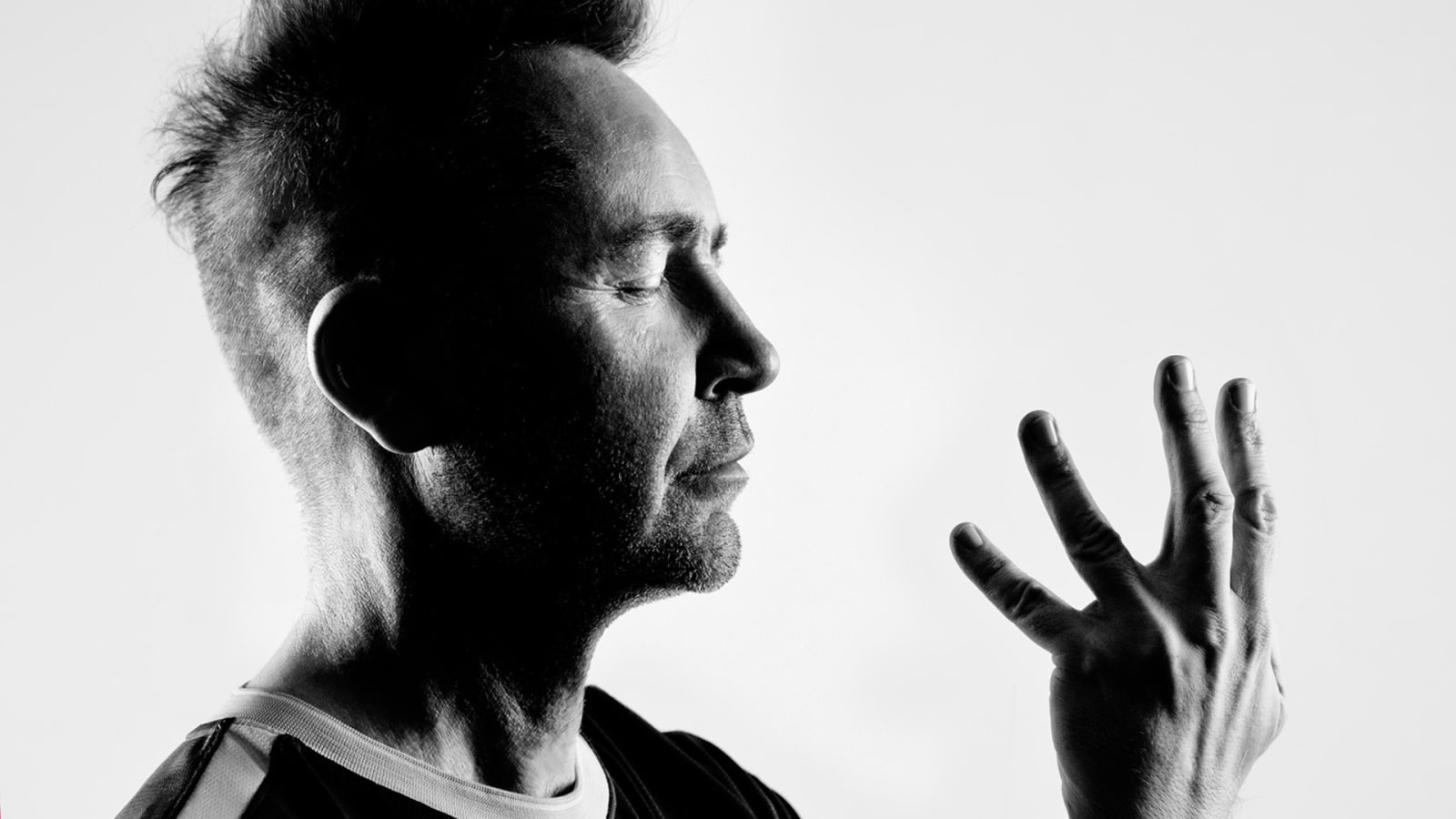 High-contrast black-and-white side profile of a person with a tall, brushed-up hairstyle, eyes closed, one hand raised with fingers spread against a pale grey background.