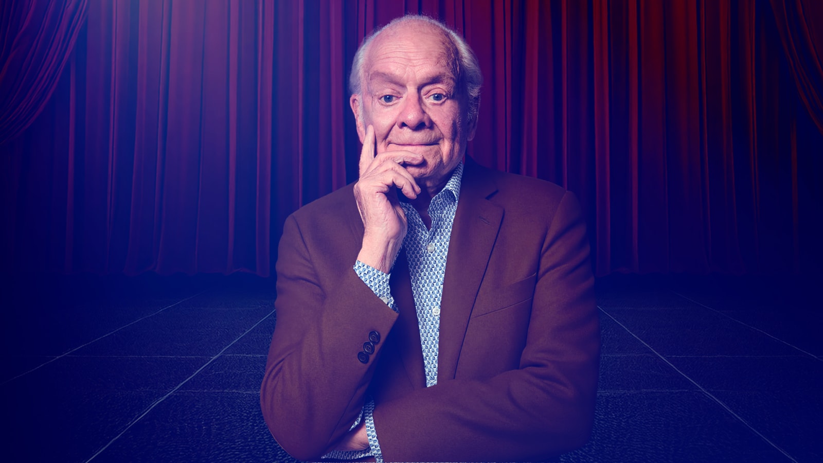 An older man in a tan blazer over a patterned blue shirt, seated with one hand raised to his chin against a deep blue and red stage-curtain backdrop