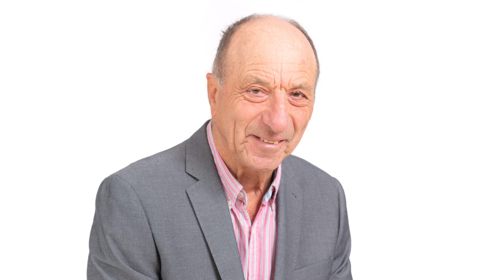 An older man in a grey blazer and open-collar pink striped shirt against a plain white background, looking slightly to one side with a faint smile.