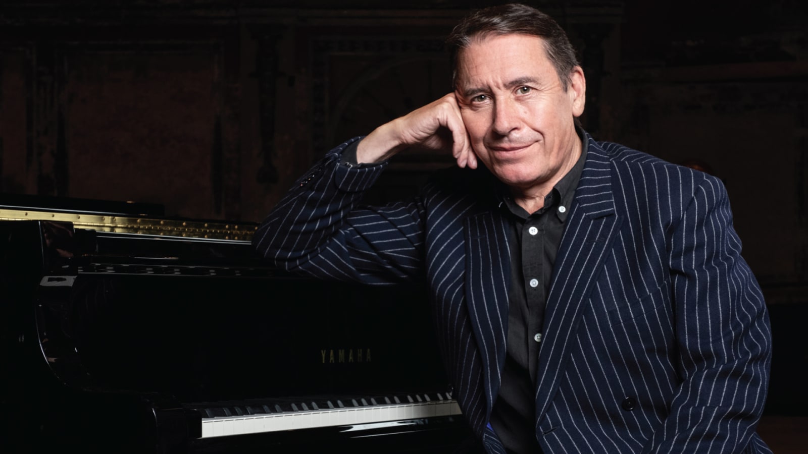 A man in a dark navy pinstripe blazer rests his elbow on the lid of a black Yamaha grand piano against a dark brick background.