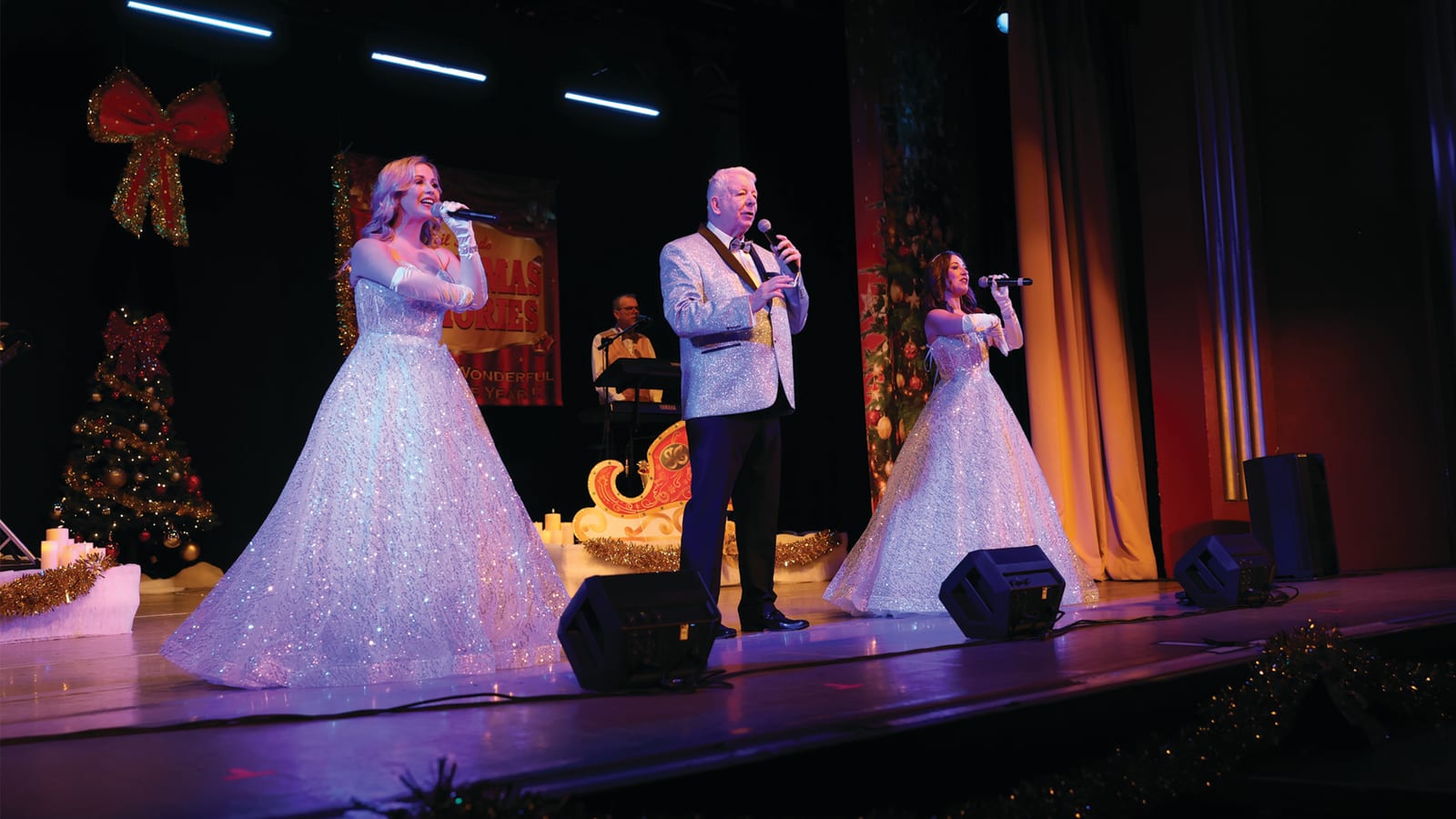 Two performers in silver sequinned gowns and a man in a silver glitter blazer on a decorated stage with a lit Christmas tree and a red sleigh prop.