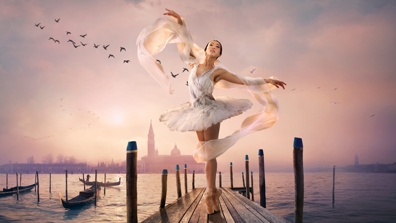 A ballet dancer in a white tutu poses on a wooden jetty with gondolas and a domed church in a soft pink dusk sky behind her.