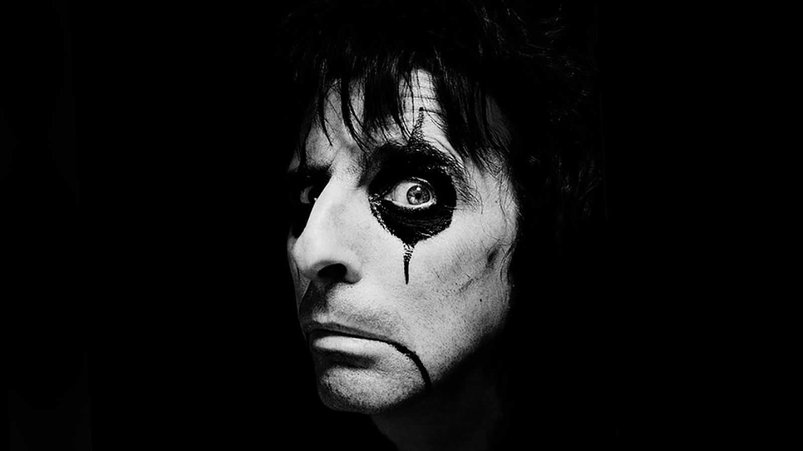 Black and white close-up portrait of a person wearing heavy black eye makeup with a dramatic drip design beneath one eye against a dark background.