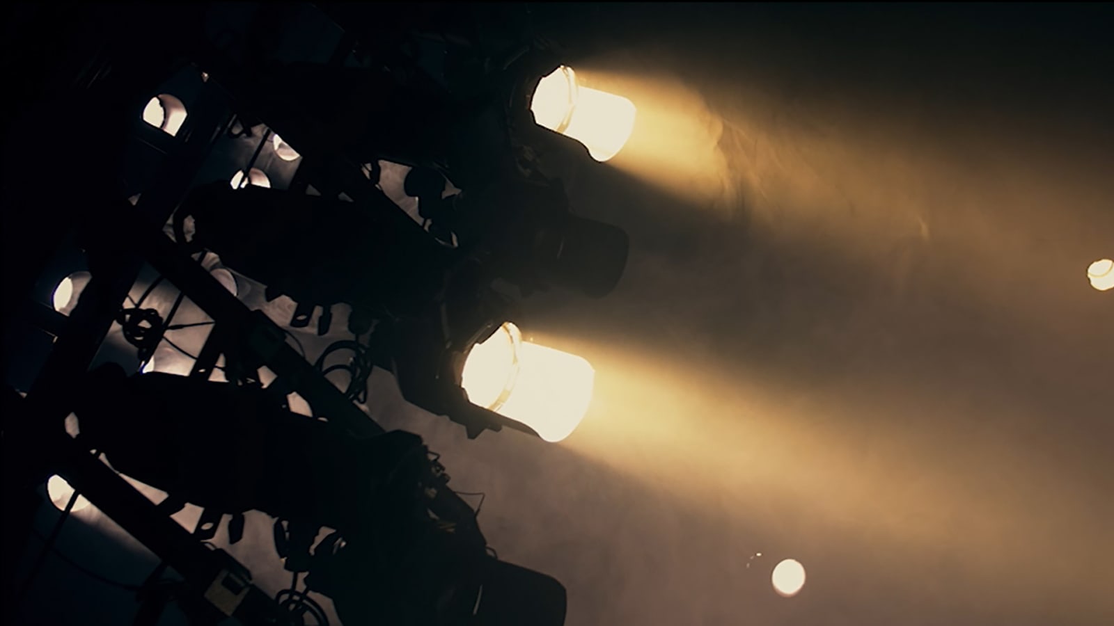Theatre lighting rig viewed from below, with two warm spotlights casting golden beams through haze against a dark background.