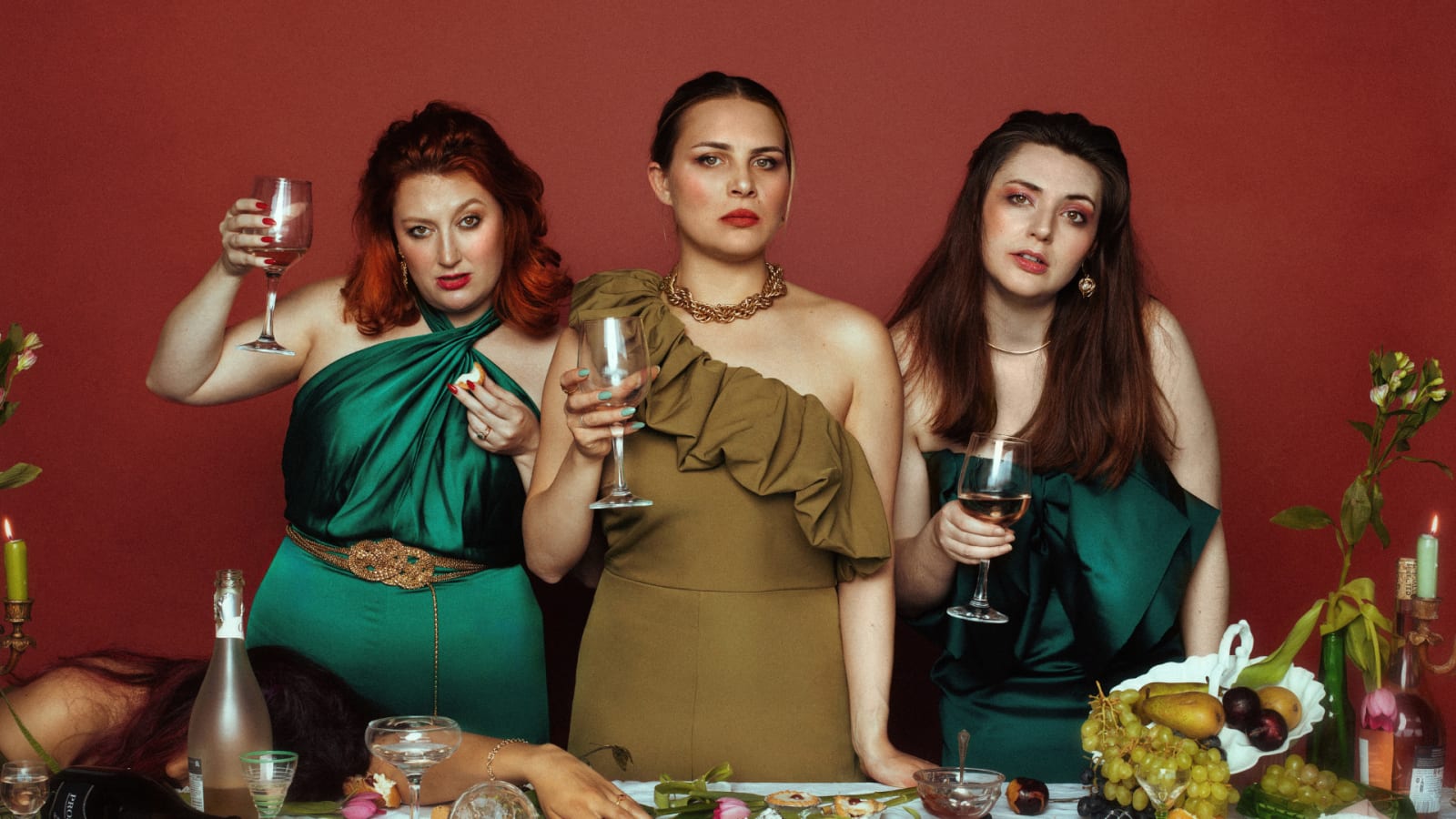 Three women in green and olive satin formal dresses hold wine glasses at a table scattered with grapes, candles and bottles against a deep red background
