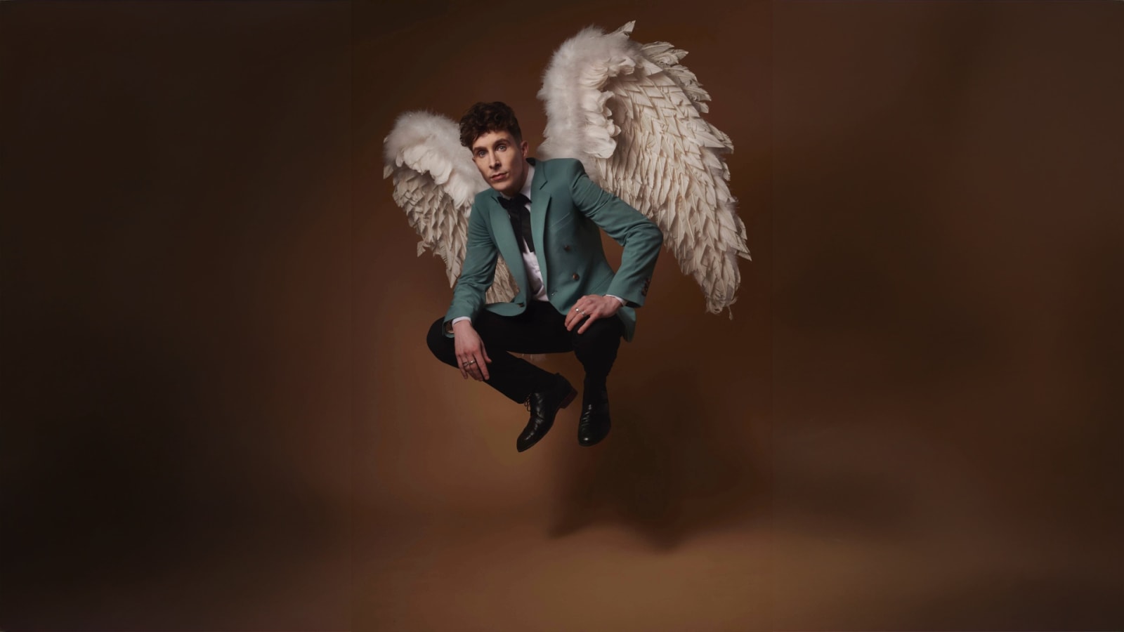 A person in a teal suit jacket and black trousers floats against a warm brown background wearing large white feathered angel wings.