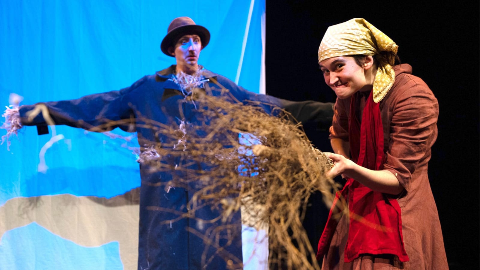 A performer in a brown dress and gold headscarf holds a bundle of hay while a second performer in a dark coat and brown hat stands behind with arms outstretched against a bright blue backdrop.