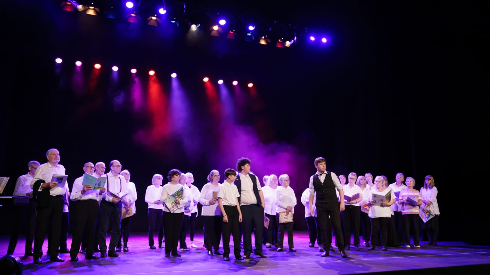 A large group of adults in white shirts and black trousers stand in a row on a darkened stage beneath red, white and purple lighting, each holding open sheet music booklets.