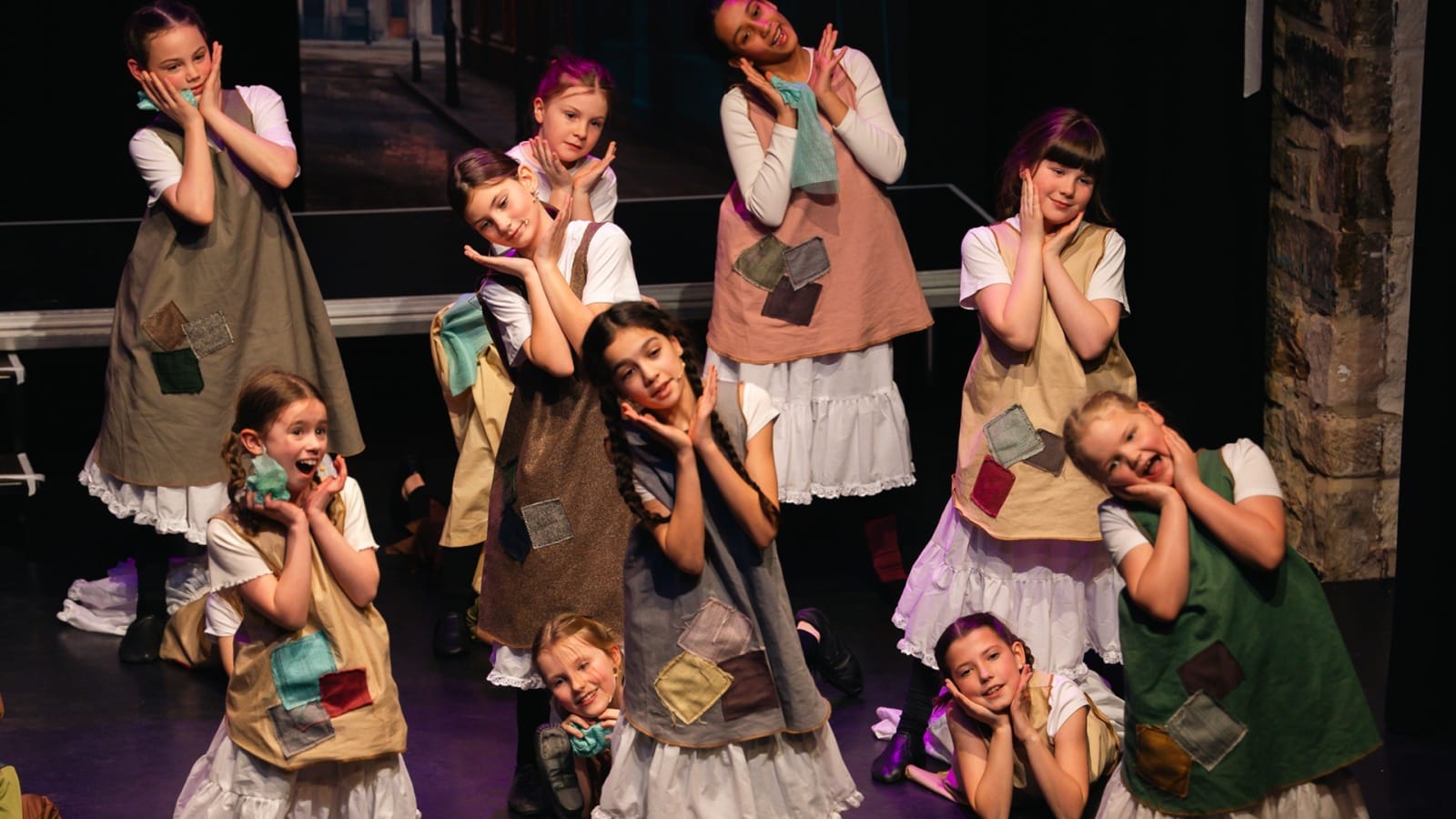 A group of young performers in patched hessian tabards over white costumes pose on a darkened stage with a cobbled street backdrop, hands raised to their cheeks.