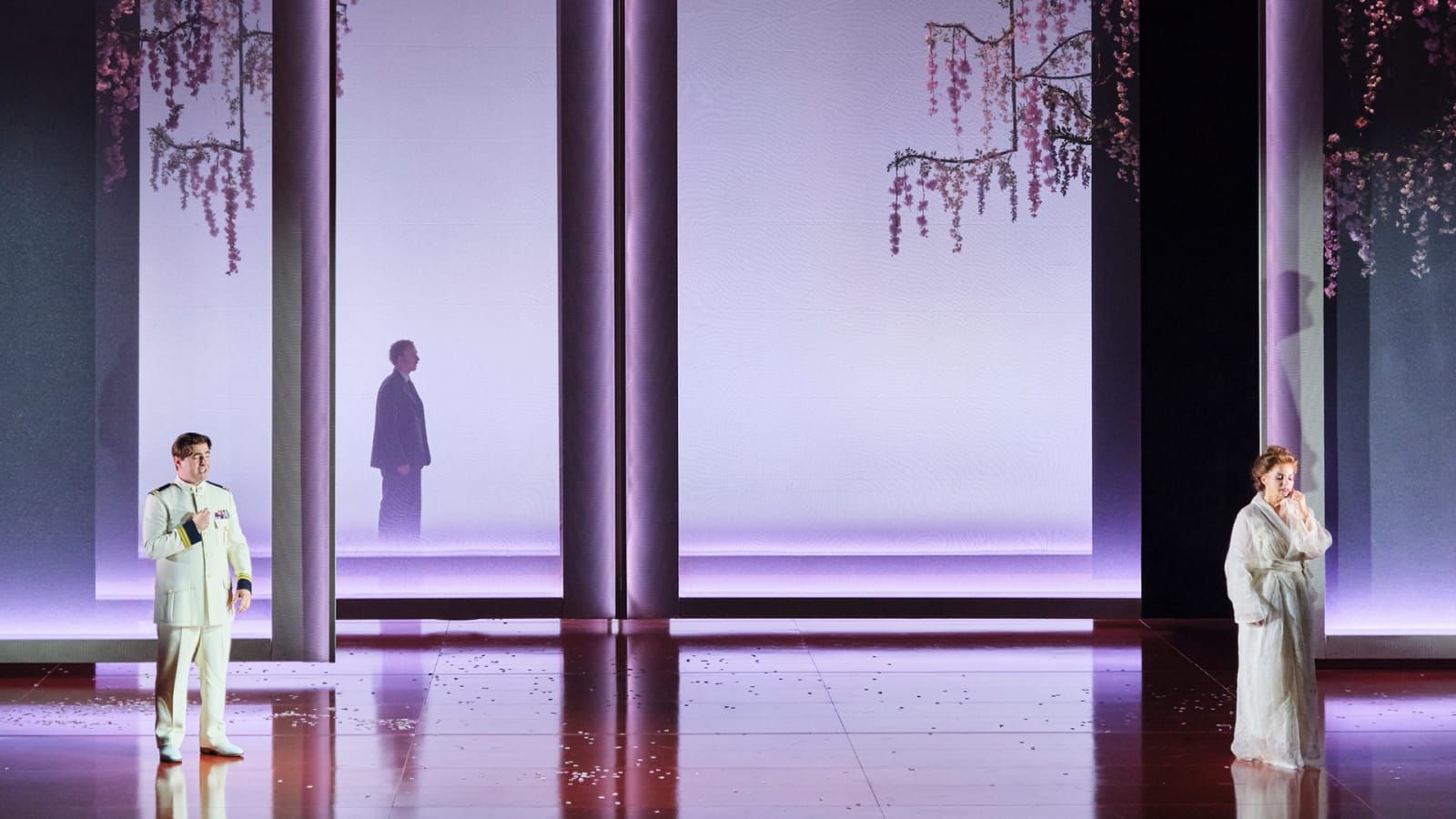 A wide stage bathed in soft purple light, with tall pale panels and hanging pink blossom branches overhead. A performer in a white naval uniform stands to the left.