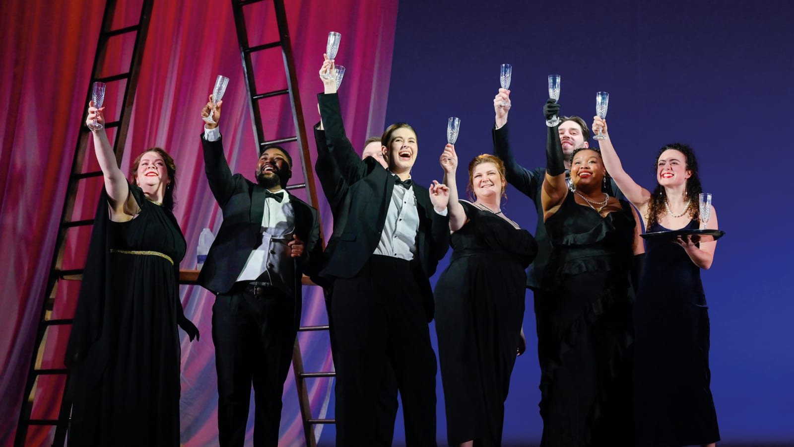 Seven performers in black formal wear raising crystal champagne flutes on a stage lit in pink and blue, with a wooden ladder visible against draped curtains.
