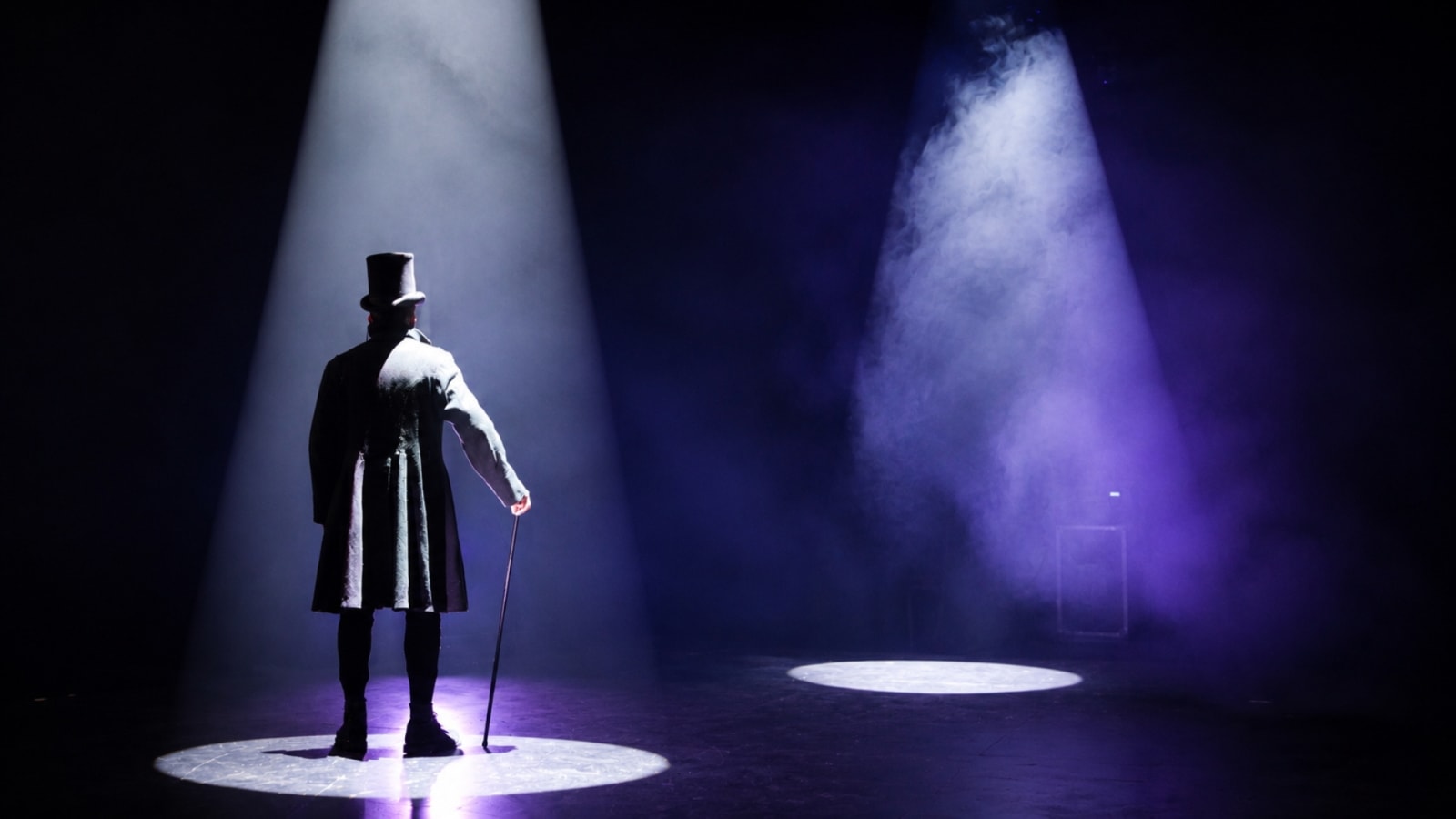 A figure in a long dark coat and top hat stands with a cane, seen from behind, lit by a single white spotlight on a dark stage with purple-tinted smoke nearby.