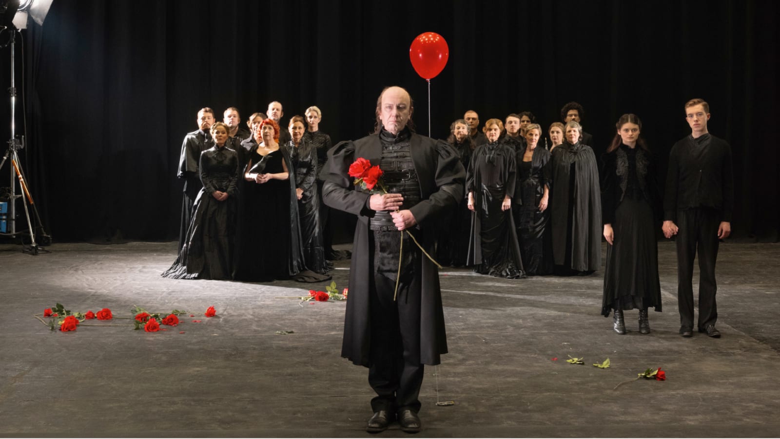 A large ensemble dressed entirely in black stands on a bare grey stage; a central figure in a long black coat holds a single red rose and a red balloon, while more red roses are scattered across the floor.