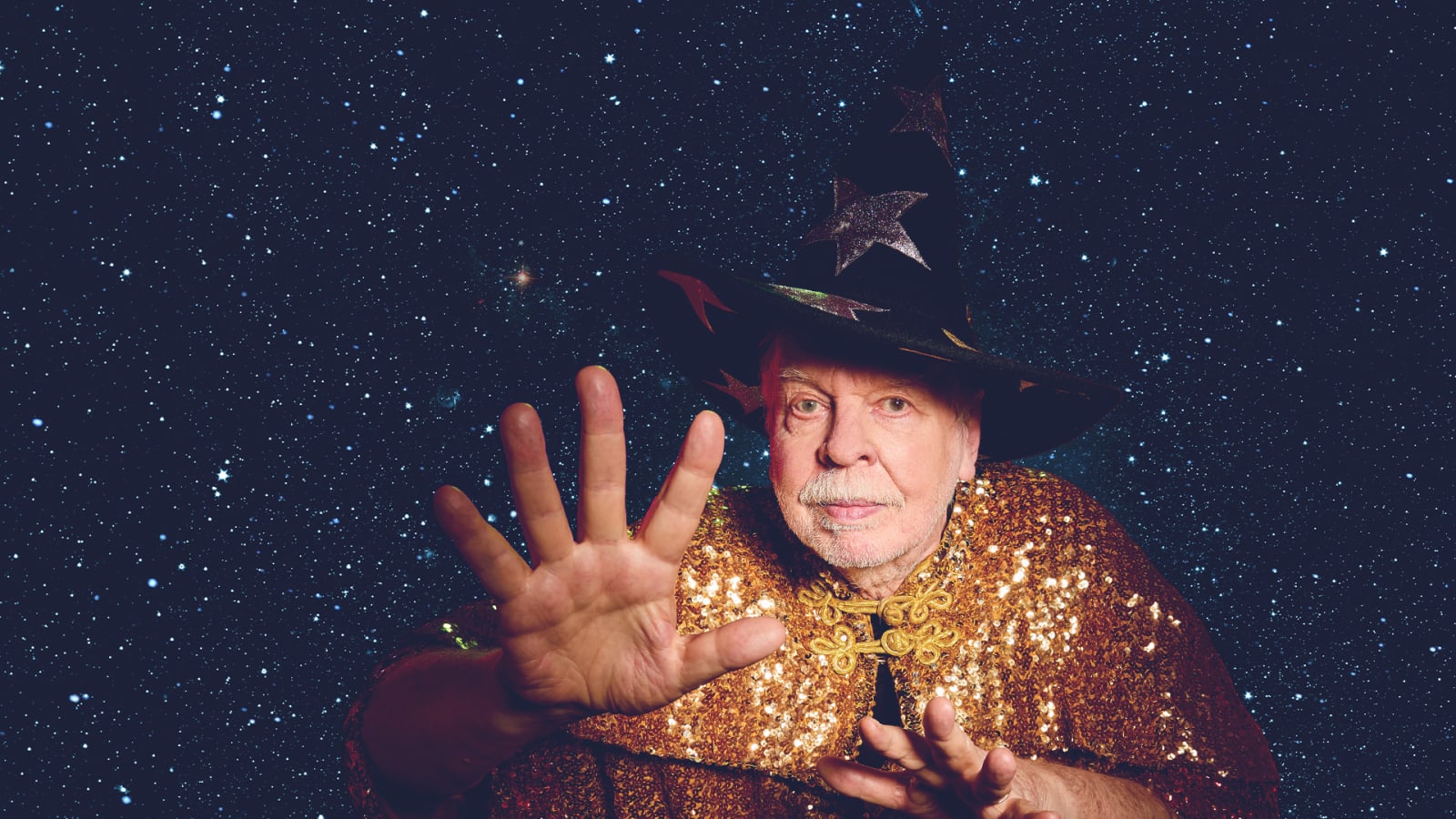 A white-haired man in a gold sequinned cape and a wide-brimmed black wizard's hat decorated with metallic stars raises one open hand towards the camera against a deep blue starfield background.
