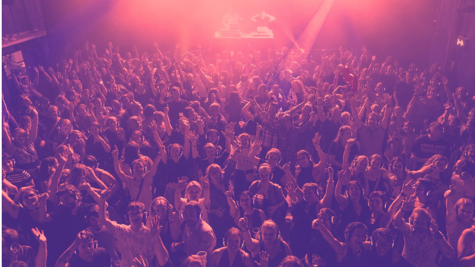 A large crowd wearing headsets with arms raised beneath pink and purple stage lighting, a DJ booth glowing at the back of the venue.