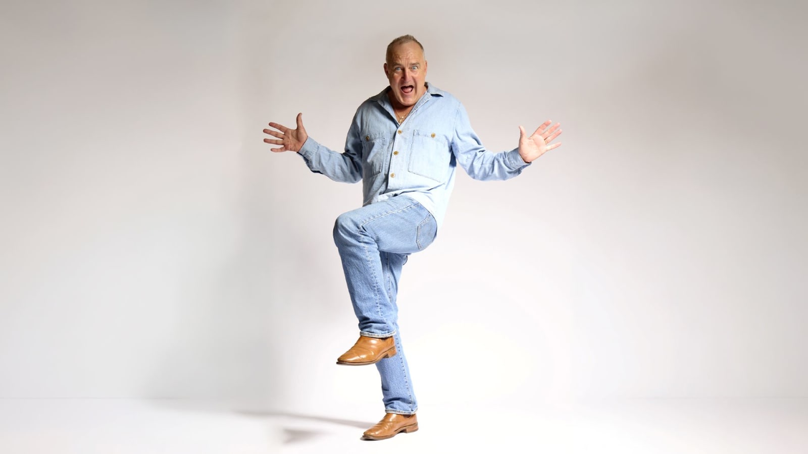 A man in a pale blue denim shirt and jeans raises one knee and spreads both arms wide against a plain white background, mouth open in an animated expression, wearing tan leather shoes