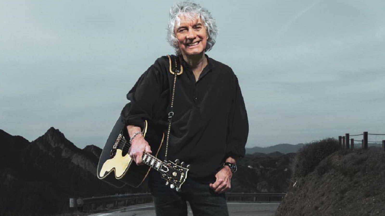 A white-haired man in a black shirt holds a black and cream semi-hollow electric guitar with a gold-trimmed strap, standing outdoors against a grey sky and dark hillside.