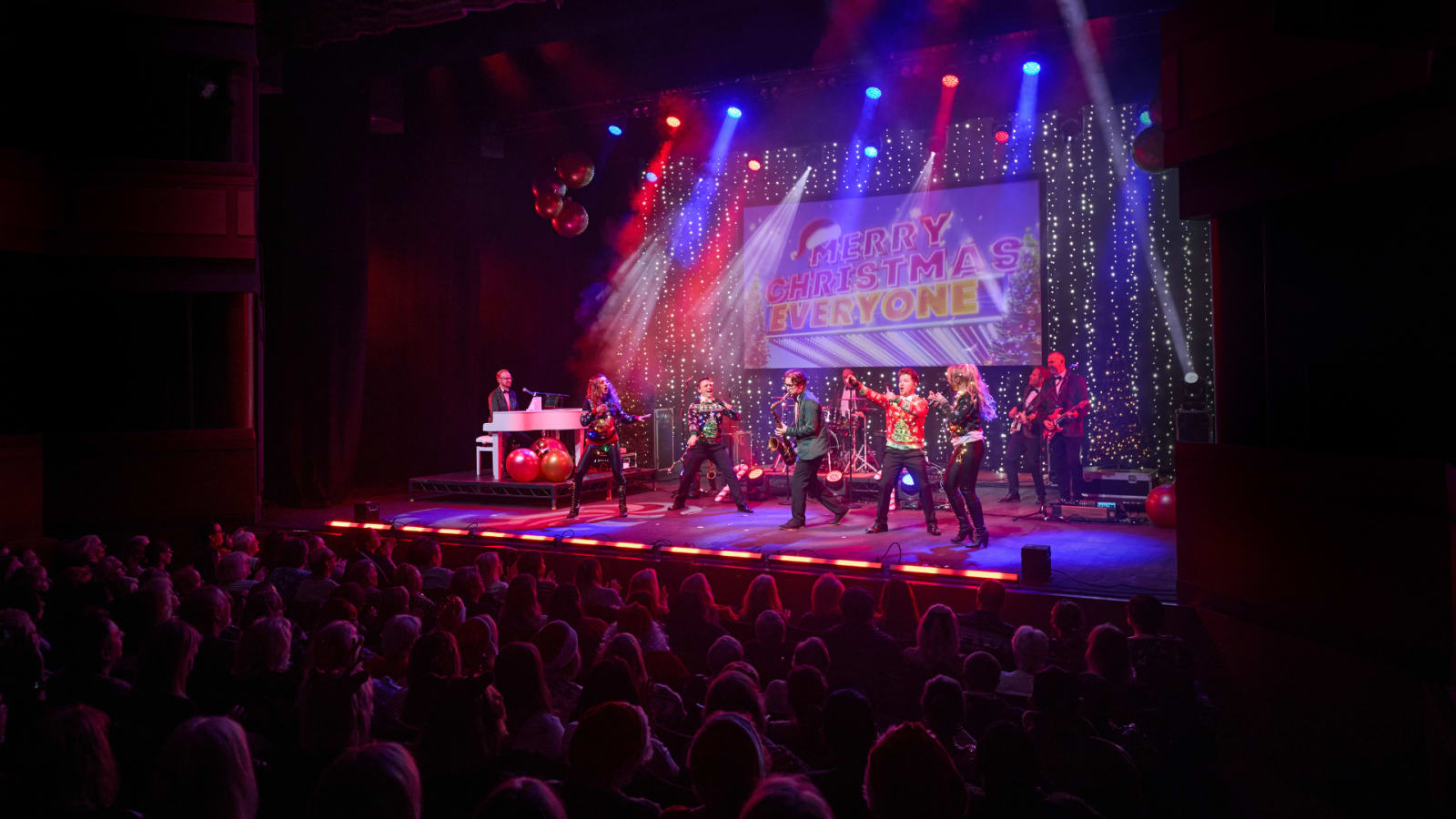 A theatre stage with red and blue spotlights, performers in festive jumpers, a screen reading 'Merry Christmas Everyone', a white piano to the left, and a full audience in the foreground.