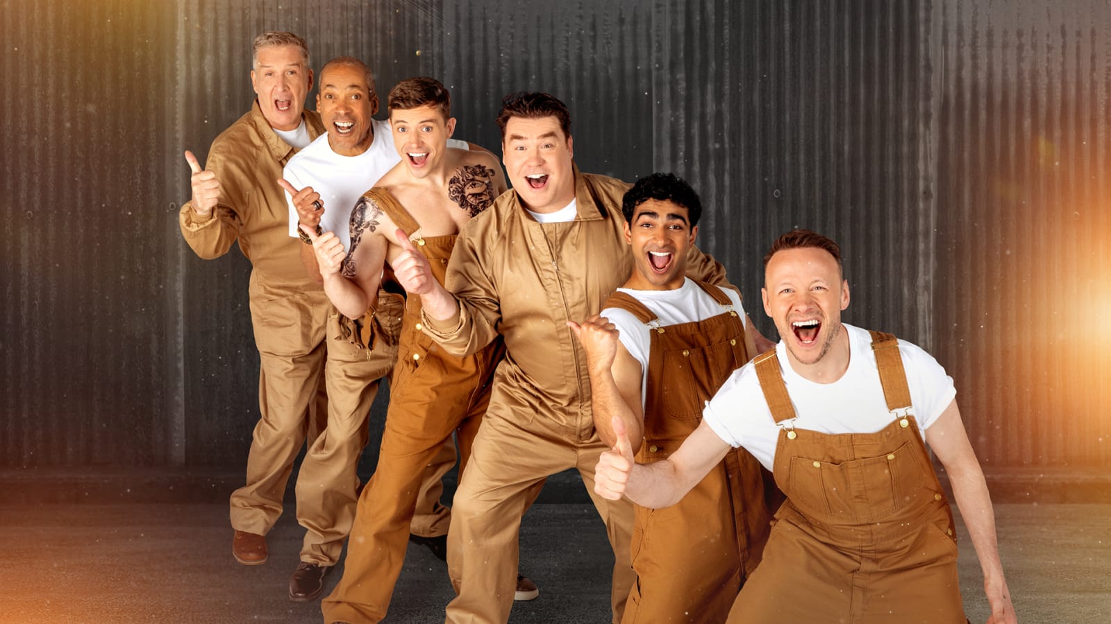 Six men in tan dungarees and white T-shirts pose in a diagonal line against a corrugated metal backdrop, grinning and giving thumbs up.