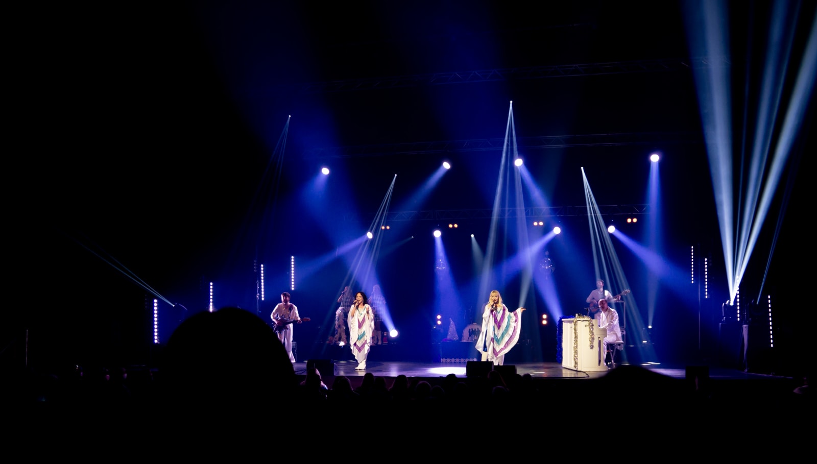 Two performers in white outfits with multicoloured chevron capes sing centre stage beneath blue spotlight beams, with band members and a silhouetted audience visible.