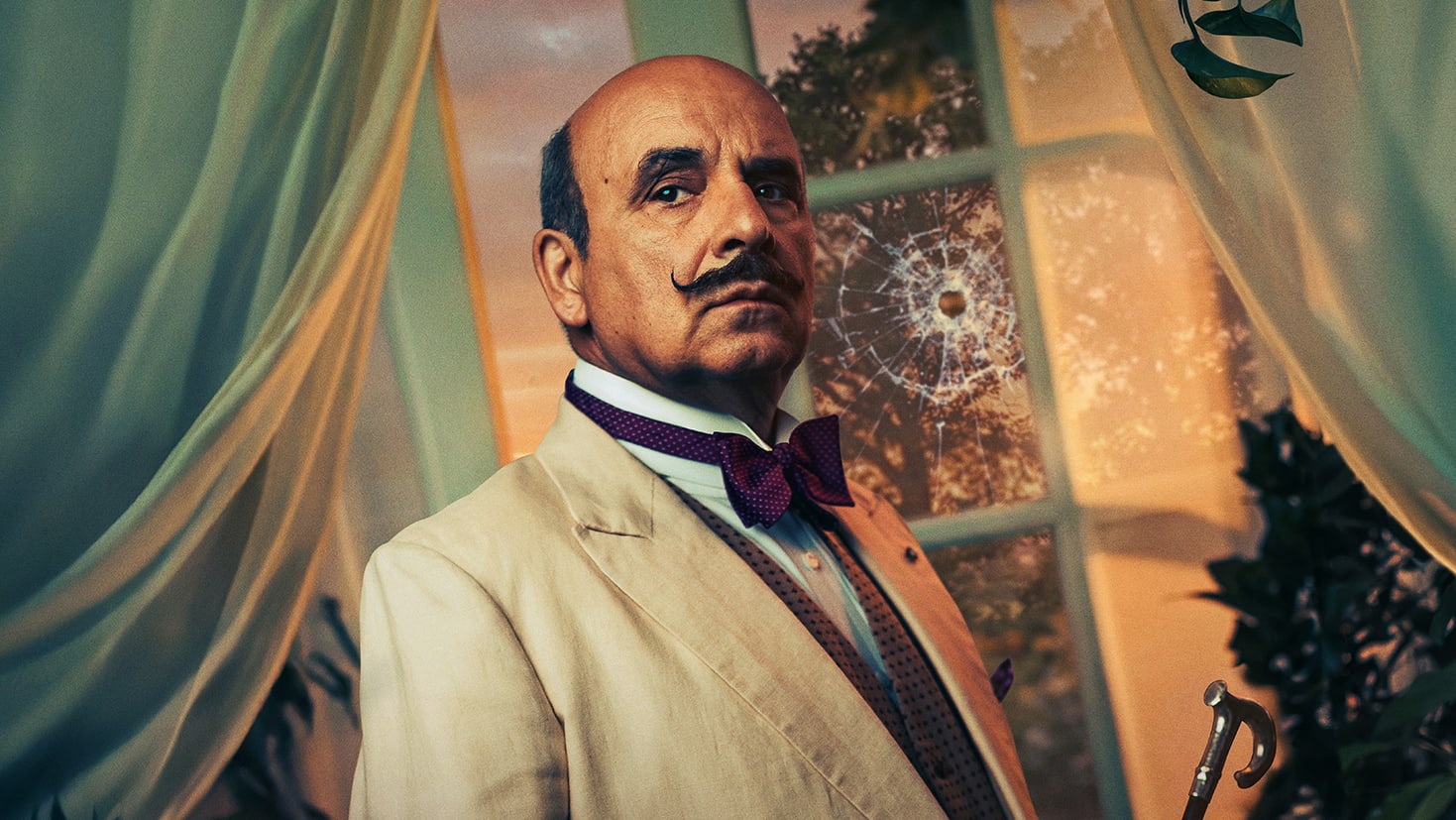A man with a waxed moustache in a cream suit and polka-dot bow tie holds a cane before a large window with a bullet hole crack and teal curtains.