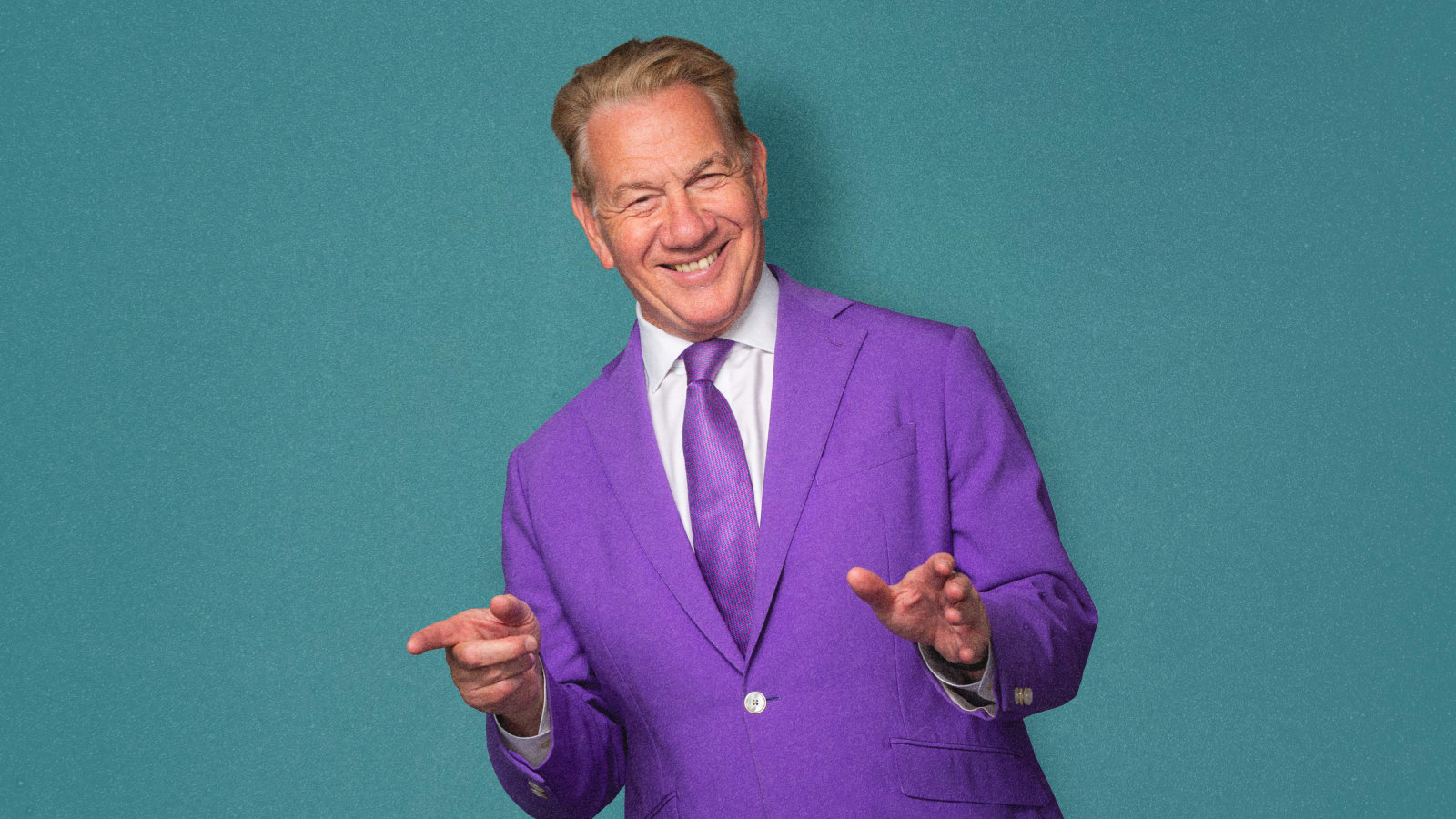 A man in a bright purple suit and tie gestures with both hands open against a teal-blue background, smiling at the camera.