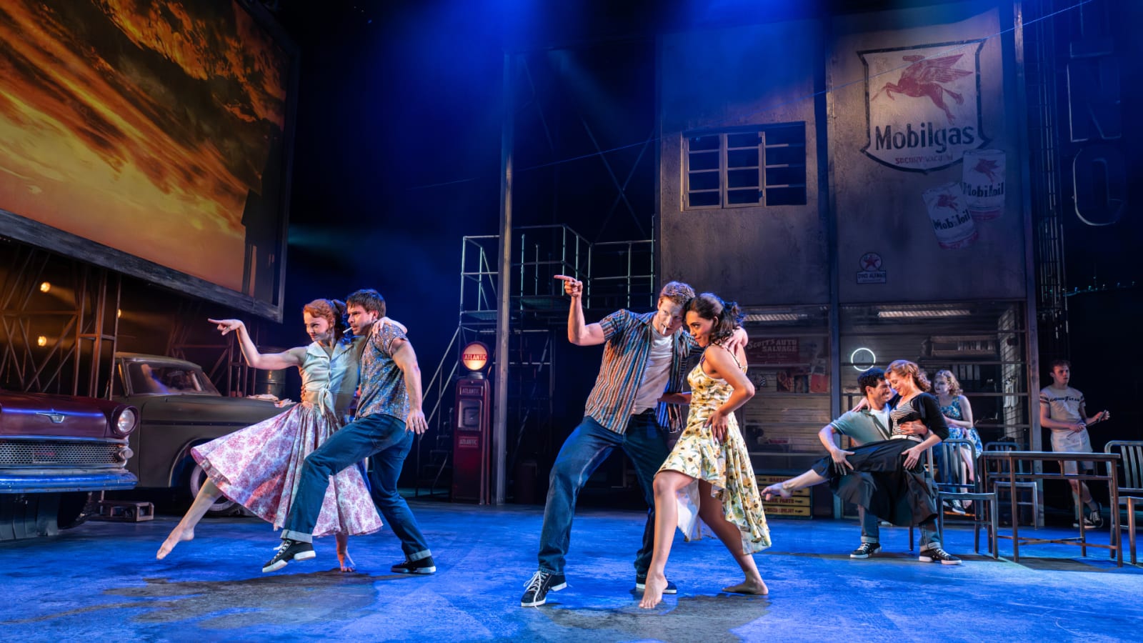 Two couples dance on a stage set as a 1950s American garage with a vintage red car, Mobilgas signage and an orange sky backdrop lit in deep blue.
