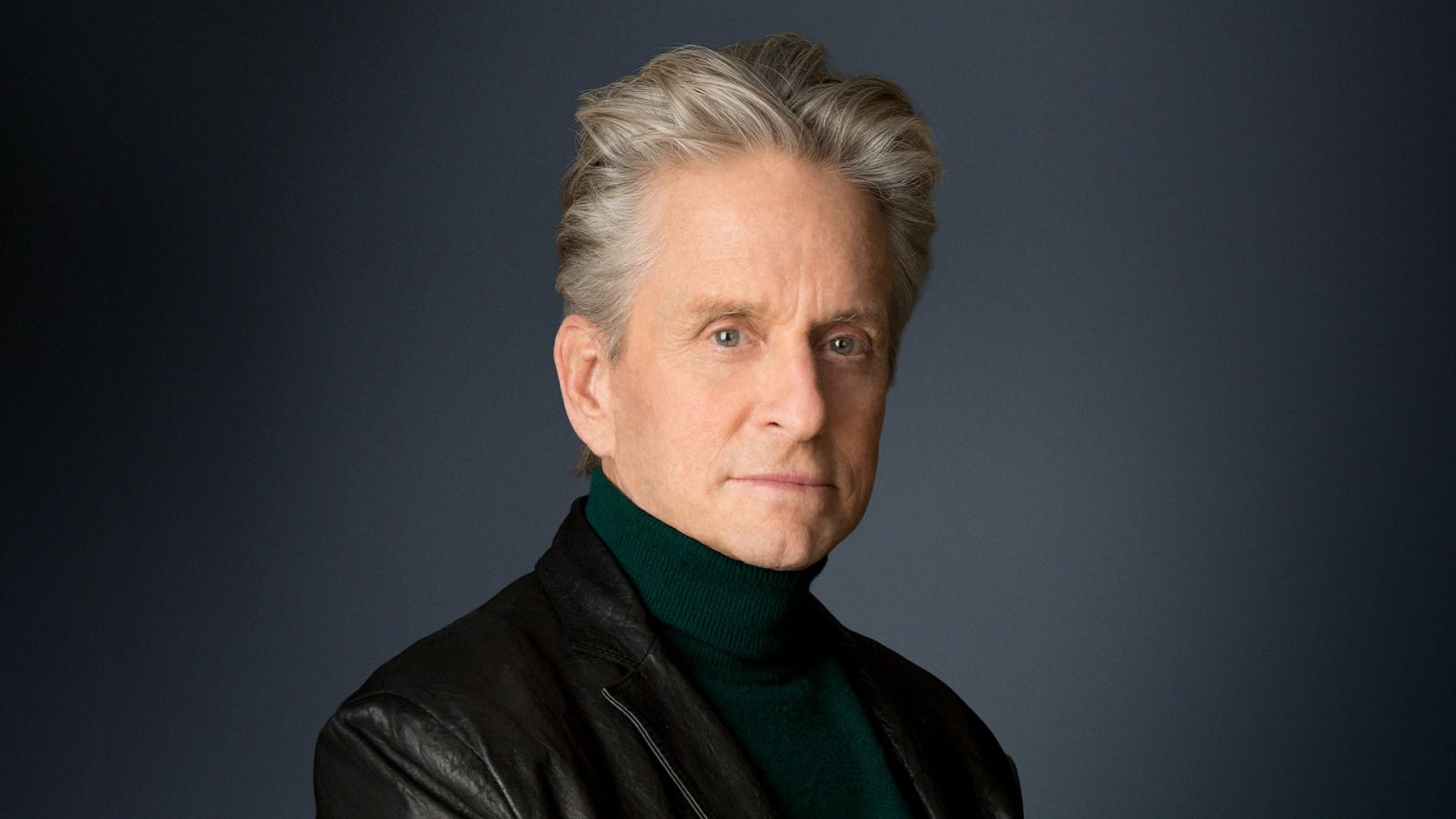 Portrait of a silver-haired man in a dark green turtleneck and black leather jacket against a dark grey background.