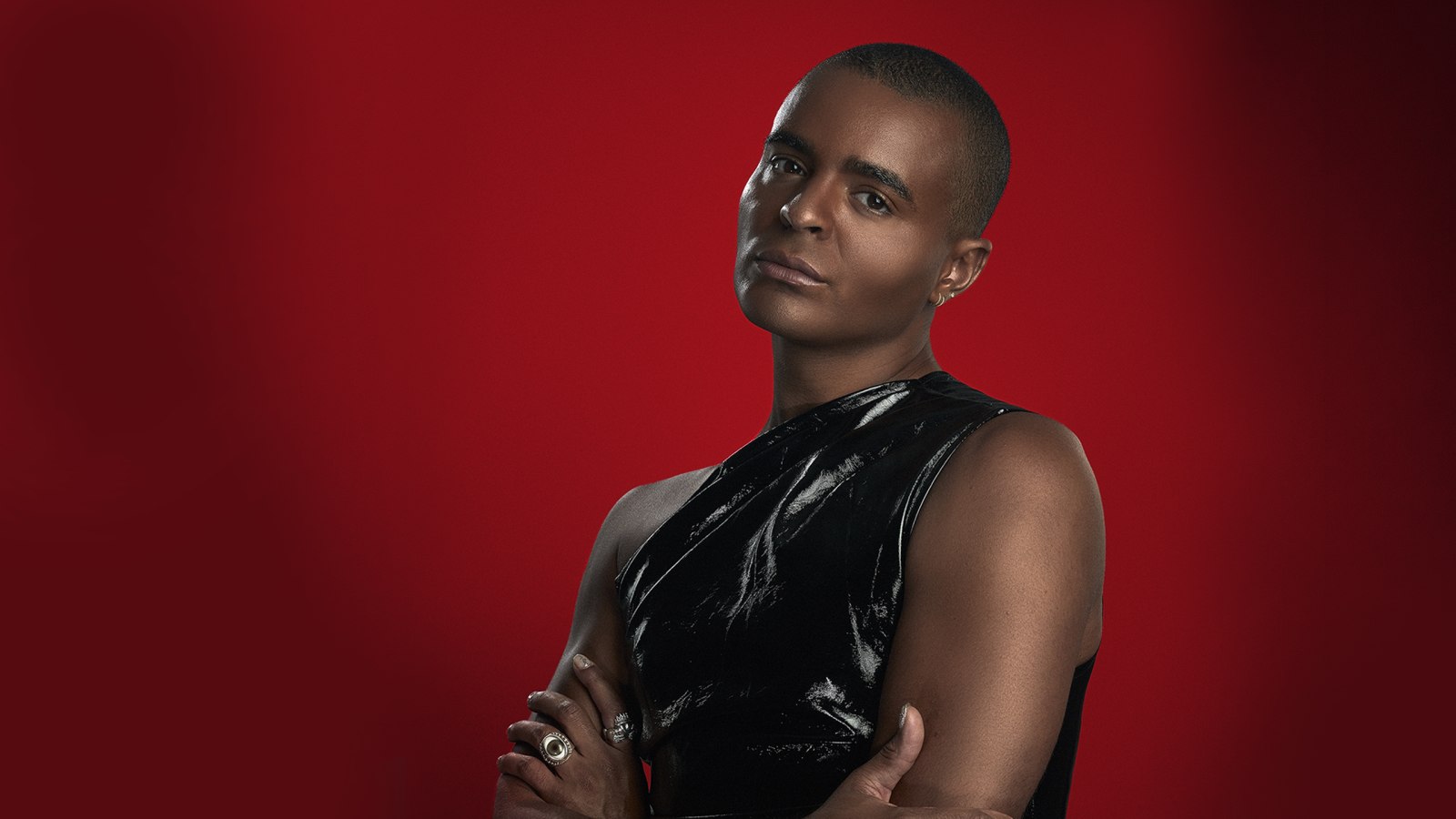 A person with a shaved head wearing a sleeveless black patent top stands with arms folded against a deep red background