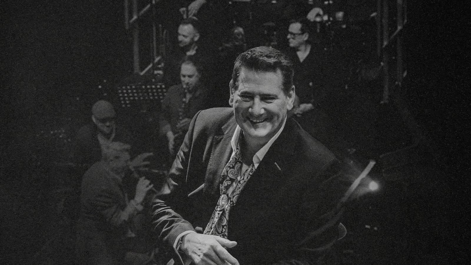 Black and white concert photograph of a man in a dark blazer and patterned shirt leaning forward and smiling, with musicians and music stands on a dimly lit stage behind him.