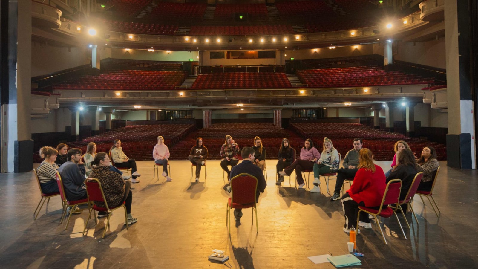 Around twenty participants seated in a circle of chairs on the bare wooden stage of a large empty theatre, with rows of red seats visible behind them.