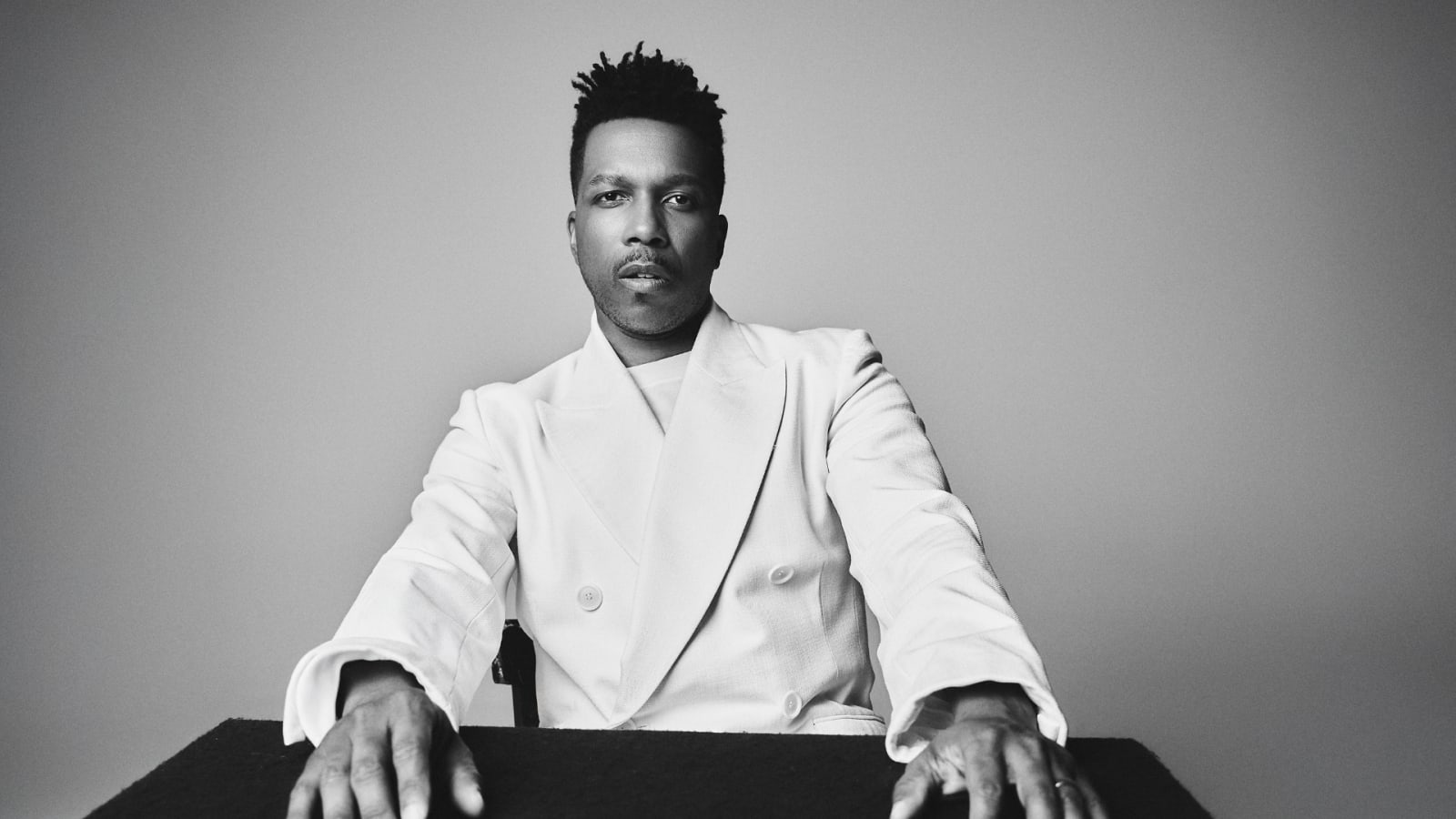 Black-and-white portrait of a man in a double-breasted white blazer, seated with both hands resting on a dark surface, looking directly at the camera against a plain grey background.