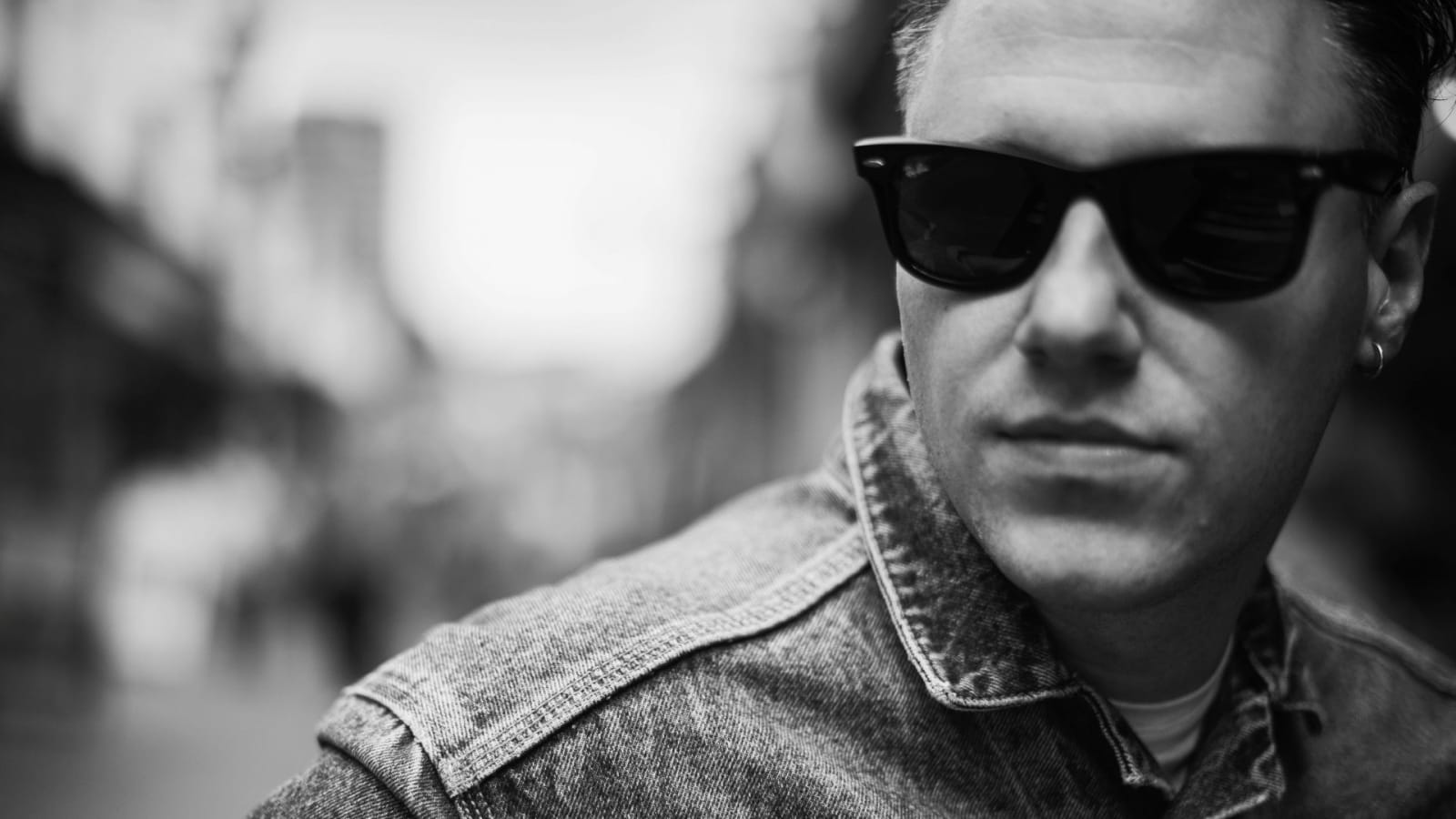 Black and white close-up portrait of a person wearing dark wayfarer sunglasses and a denim jacket, with a blurred outdoor background