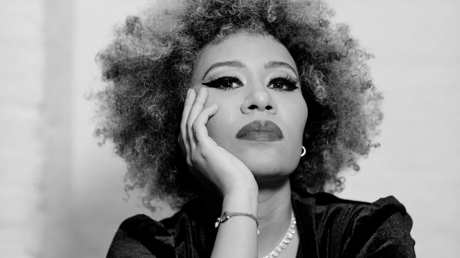 Black and white close-up portrait of a person with a voluminous curly afro, bold winged eyeliner, pearl necklace and velvet top, resting their cheek on one hand and gazing upward