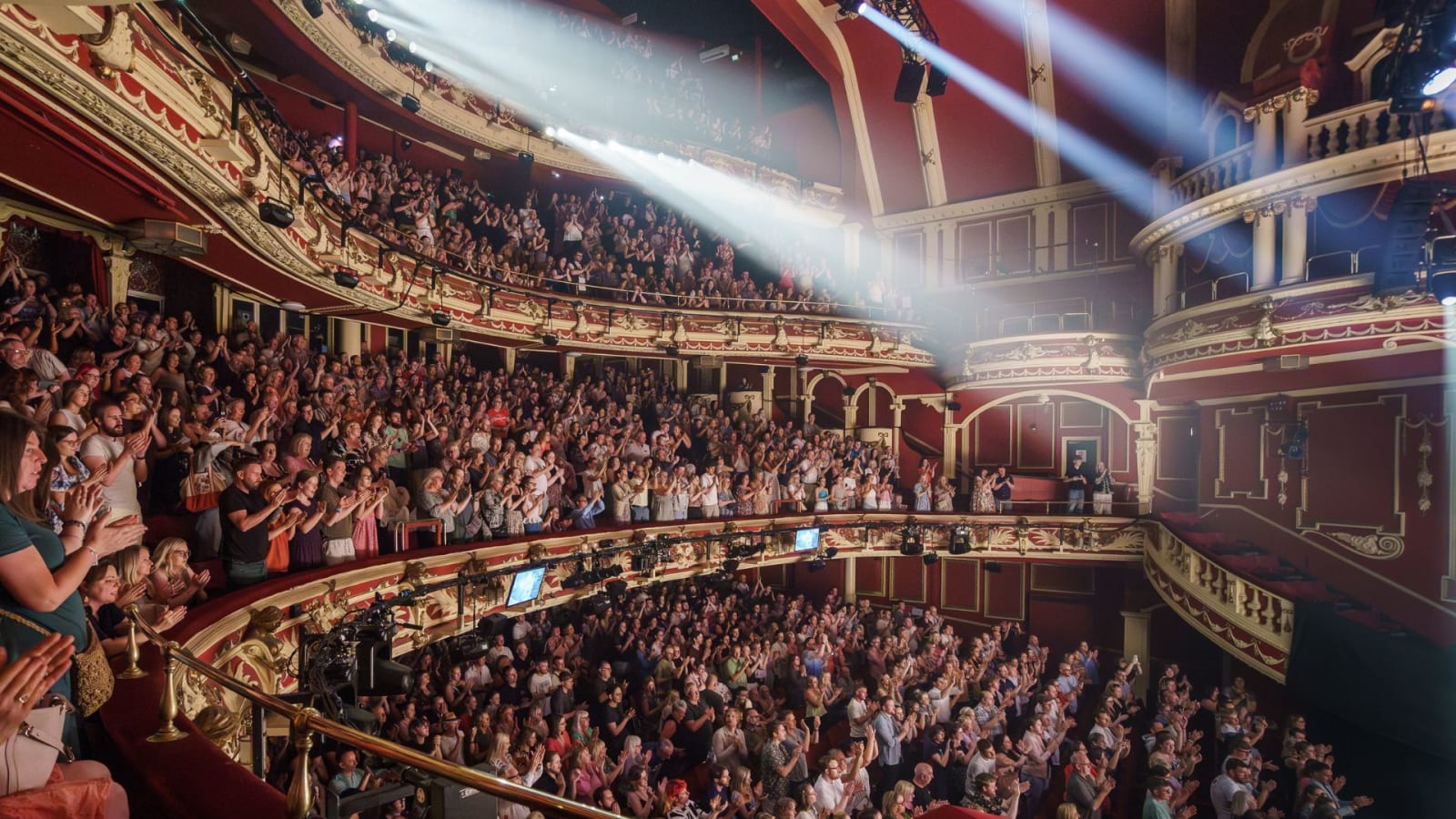 A packed theatre auditorium with ornate red and gold balconies and a standing audience under bright stage spotlights.