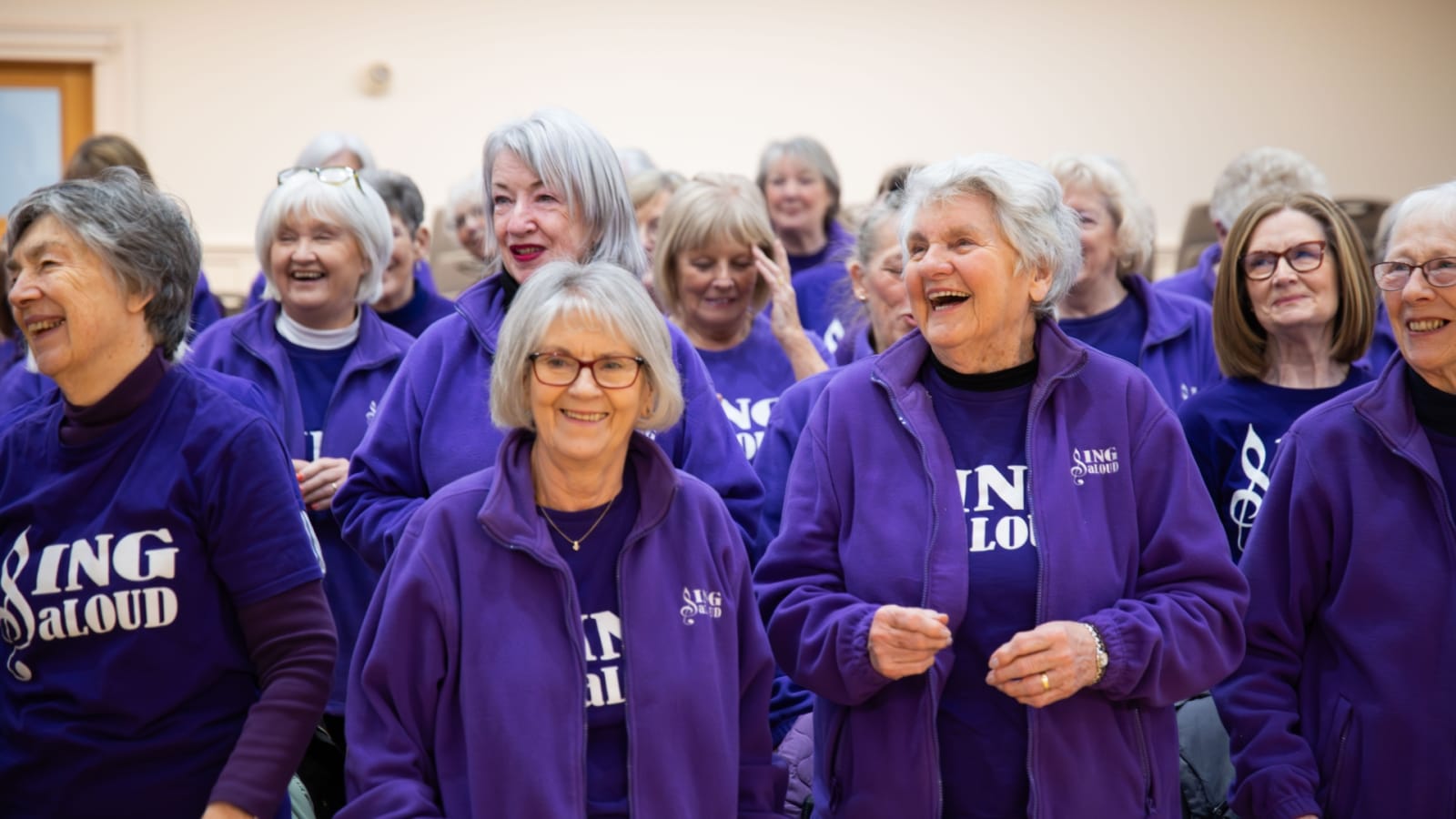 A large group of choir members wearing matching purple zip-up fleeces and T-shirts bearing the Sing aLoud logo, gathered together in a bright rehearsal room