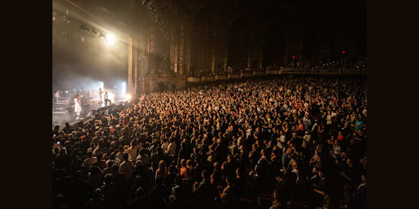 A wide crowd shot of a full audience in the general admission pit at Kings Theatre. The audience is illuminated by the stage lights as a band is performing on stage.