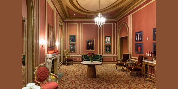 A shot of one of the premium lounges inside Kings Theatre. It is an historic room with an ornate carpet, lush furniture, a fireplace, and framed photos of past performances.