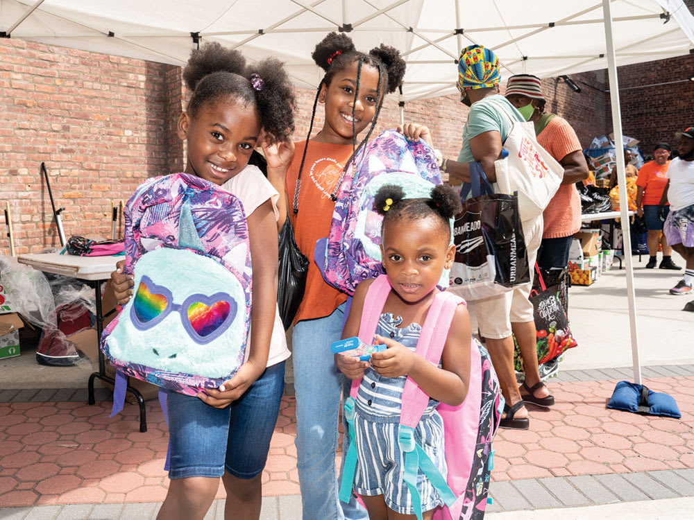 A shot of three Black girls smiling to the camera, holding up their new free colorful backpacks they received at the annual KingsCares Back to School event in the courtyard next to the theatre.