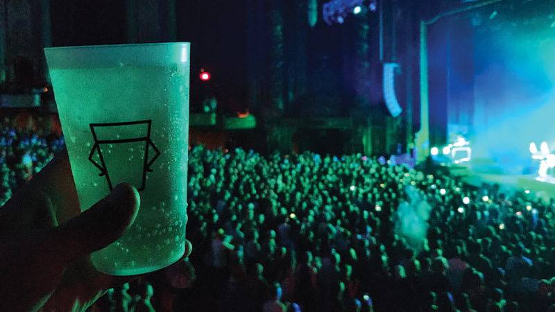 An up-close shot of a recyclable Cup Zero cup. A person is holding it in the foreground, with a live concert happening in the background.