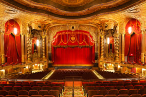 A brightly lit shot of the house inside Kings Theatre. The view is from the mezzanine and features the orchestra section and the stage with the curtains drawn. The inside is a historic and ornate renovated theatre.