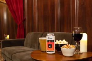 A still life shot of beer and wine in glasses, and various snacks in bowls on a side table. A couch can be seen in the background with wood paneled walls behind it. This is in one the premium Speakeasy lounges at Kings Theatre.