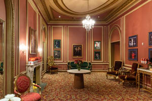 A shot of one of the premium lounges inside Kings Theatre. It is an historic room with an ornate carpet, lush furniture, a fireplace, and framed photos of past performances.