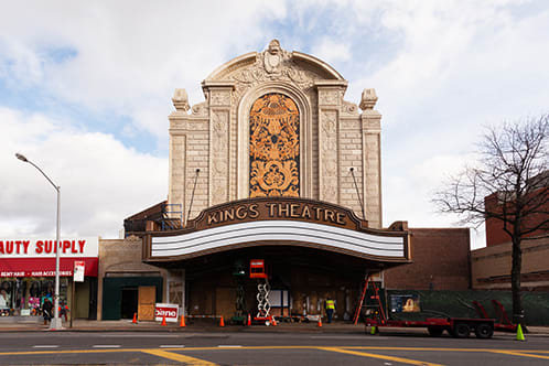 An exterior shot of Kings Theatre from across the street on Flatbush Ave. Kings Theatre's exterior is an ornate old theatre with a central marquee out front.