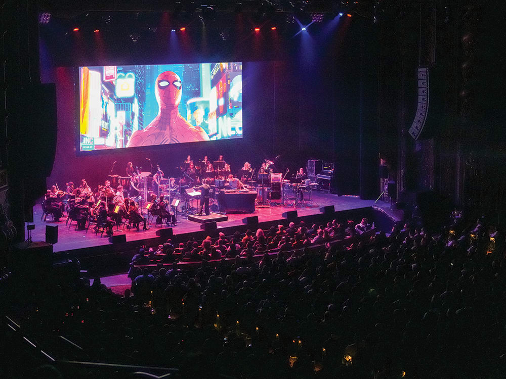 A shot of an orchestra on stage with a crowd watching at Kings Theatre. Behind the orchestra is a large screen with Spiderman on it.